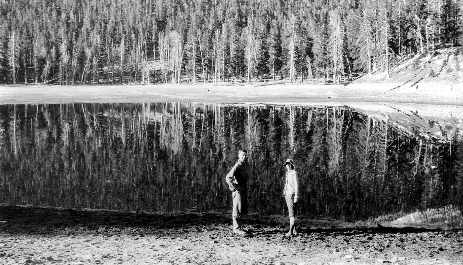 Two hikers standing on the sandy shore of Lake Cuyamaca with pristine, mirror-like reflections of tall pines across the water.