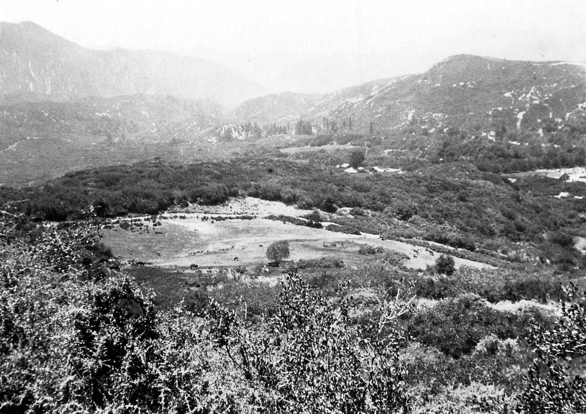 An expansive view looking down from a high ridge across a vast, rolling wilderness valley filled with dense brush and distant cabins.