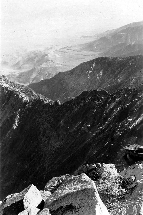 A dramatic, high-altitude vista looking across steep, rocky mountain ridges toward towering Sierra Nevada peaks in the distance.