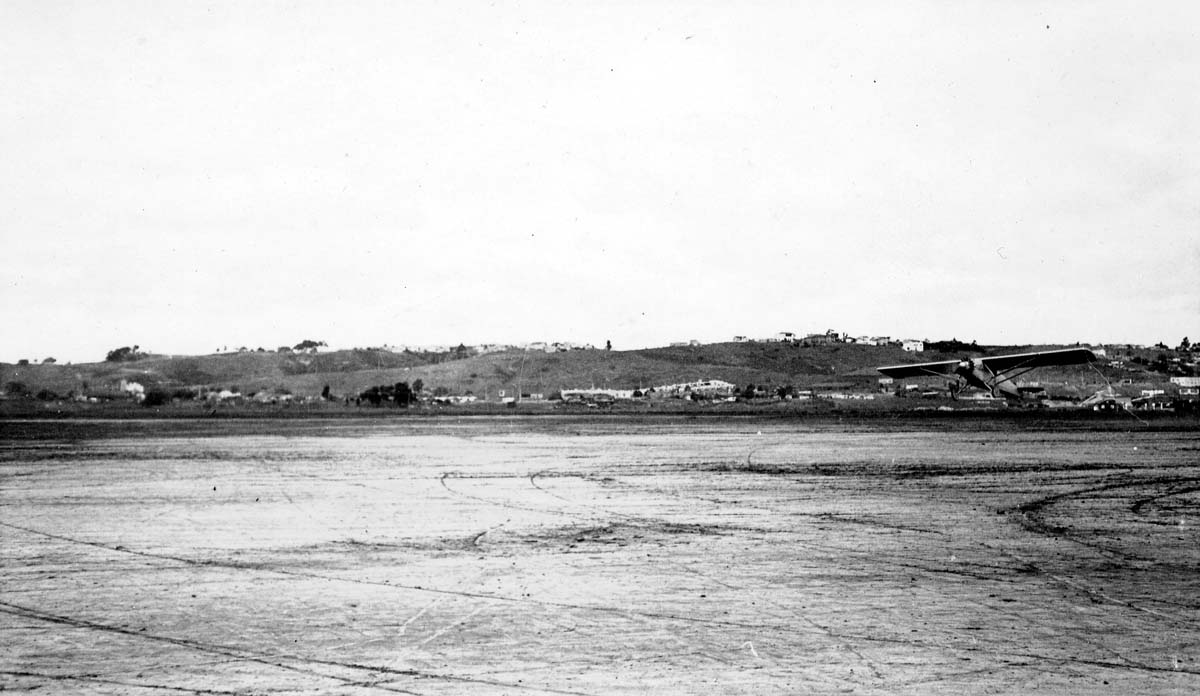 Long-distance airfield photograph of the Spirit of St. Louis airborne low in ground effect across Dutch Flats.