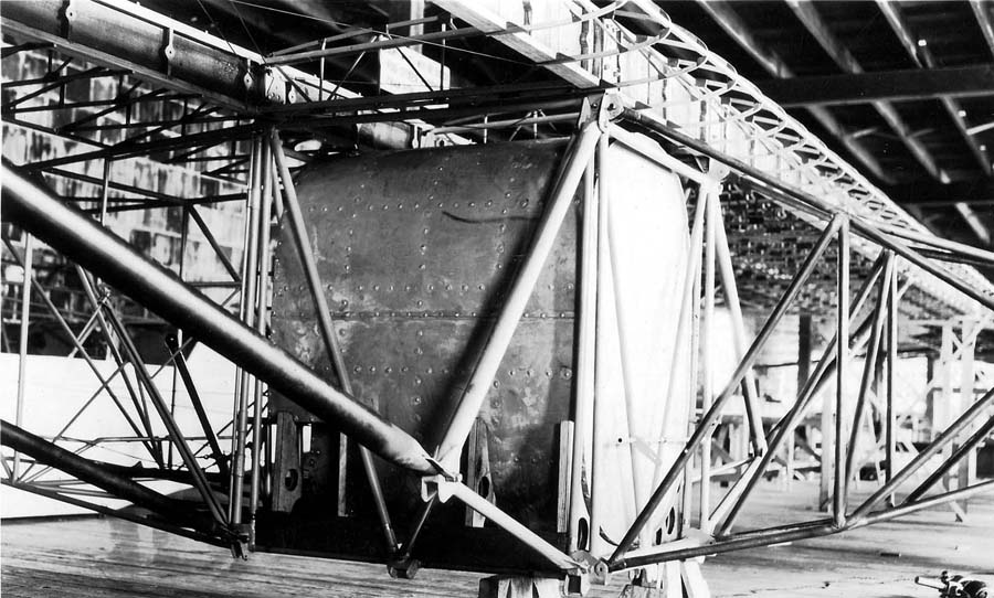 Factory view of uncovered fuselage frame, main fuel tank rivets, and wooden wing structure on sawhorses.
