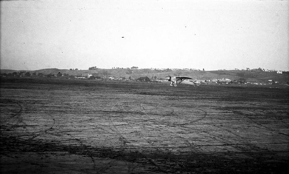 Telephoto airfield photograph of the Spirit of St. Louis flying level at low altitude across Dutch Flats.