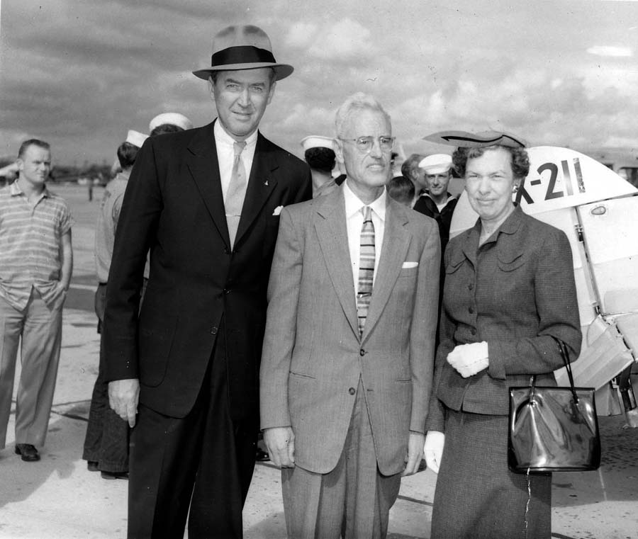 A vintage black and white photograph of actor James Stewart and Spirit of St. Louis designer Donald A. Hall and wife Elisabeth Hall in-front of a aircraft replica in California.