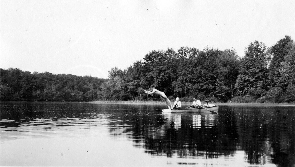 Vintage Black and White photograph of male diver captured in mid-air as jumps from a boat, into a lake with three others in the boat. Photo taken by Donald Hall.