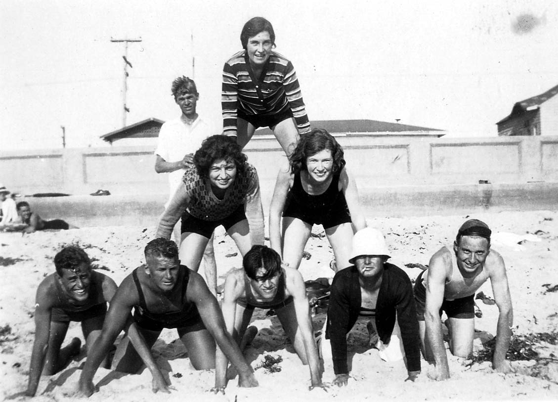Funny vintage photo of Donald Hall smiling at the camera with eight other young adults balanced in a tower on the beach in Pacific Beach, California. Hall is in the lower left corner.