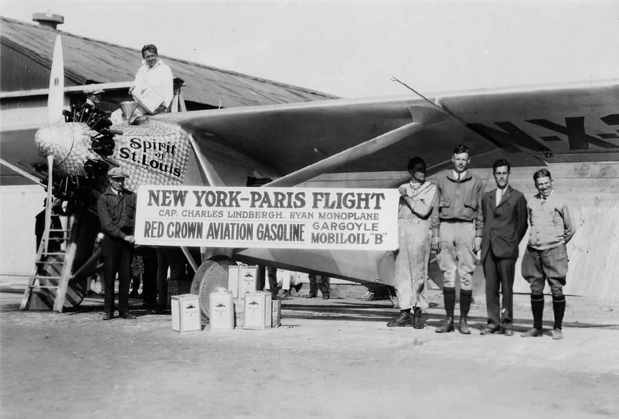A vintage black and white photograph of the Spirit of St. Louis with sales manager AJ Edwards, engineer Donald A. Hall, pilot Charles Lindbergh and other workers holding up a promotional sign for Red Crown aviation fuel and oil after the plane was finished in May 1927 at Dutch Flats airfield, San Diego, California. A man is sitting on the plane, pouring fuel into the aircraft.