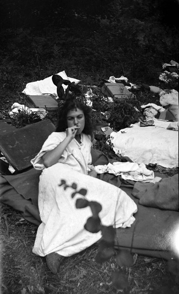 A vintage photograph of a female smoking a cigarette in a light colored dress in a wooded area. She is laying down on a bed of blankets with various luggage and other clothes on the ground.