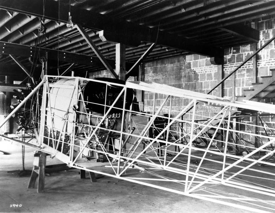 A vintage black and white photograph of the Spirit of St. Louis during assembly in the factory with the newly attached Wright J-5c Whirlwind engine. The main fuel tank, control panel, and uncovered fuselage is visible. Photo was taken by Chief Engineer Donald A. Hall, in San Diego in May 1927.